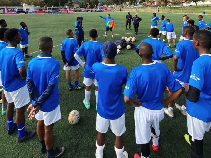 Young footballers in blue jerseys gather around a coach as part of Espanyol Academy SA’s 2025 player development camp.
