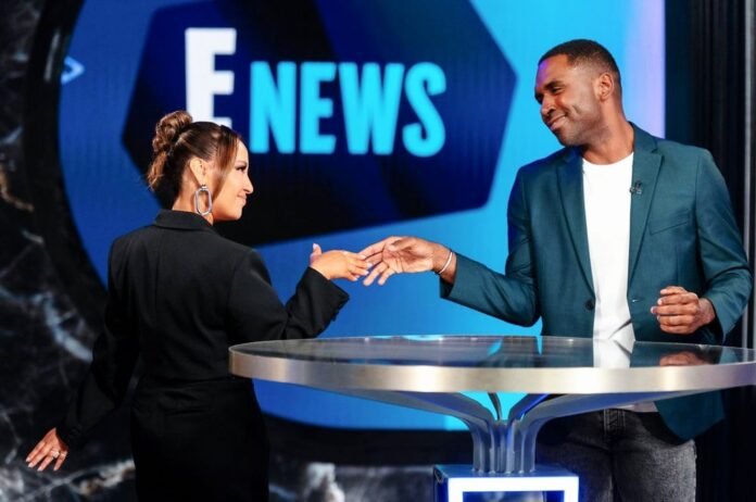 Smiling E! News hosts on set holding hands.