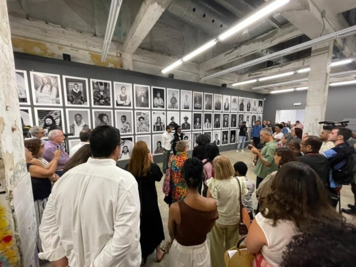 A wide shot of a crowded art gallery with an industrial ceiling, featuring a massive wall of black and white portrait photography.