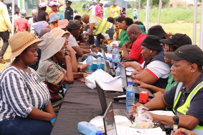SASSA staff assisting SRD grant beneficiaries at a community registration and verification centre, with residents seated across tables using laptops to check grant status and resolve payment issues.