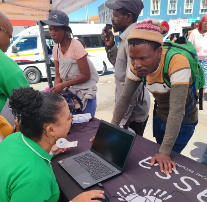 SASSA official helps young man with SRD grant at outdoor service desk, others wait in line.