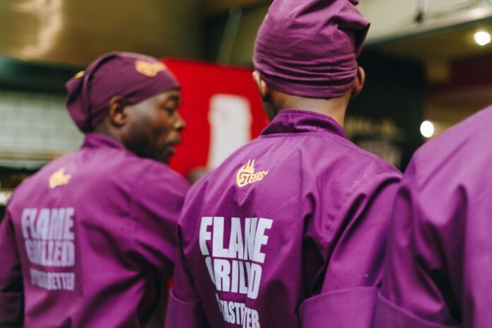 Three Steers employees in purple chef jackets and headwraps with “FLAME-GRILLED IT TASTES BETTER” text, photographed in a kitchen environment.