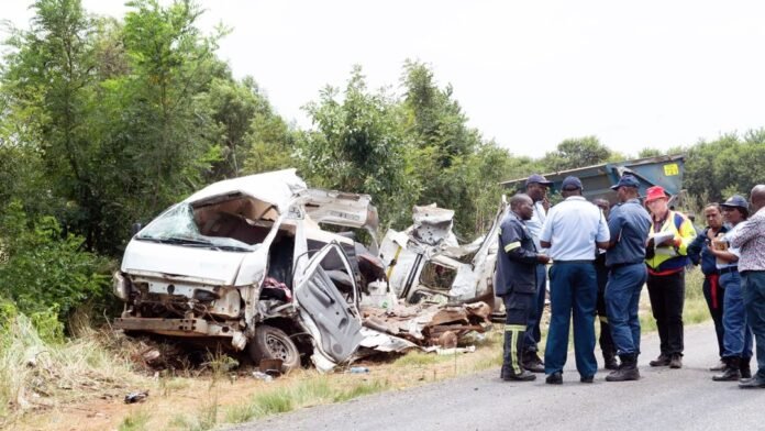 The severely mangled wreckage of a white scholar transport minibus rests on a grassy embankment next to a road in Vanderbijlpark, with police officers and emergency responders standing nearby conducting investigations.