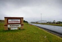 Minister signs off: East London becomes KuGompo City in landmark decision A brick-based roadside welcome sign is on a grassy verge next to a wet asphalt road under an overcast, grey sky. The top portion of the sign features the words "Welcome to" followed by "East London," which has been struck through with a thick red line. Below this, a newer, clean white panel has been installed featuring the name "KuGompo" in bold black lettering. In the background, a long industrial-style building and streetlights stretch into the distance along the roadside. Roadside welcome sign showing “East London” crossed out in red and replaced with “KuGompo City” on a new panel beside a highway under cloudy skies.