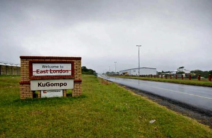 A brick-based roadside welcome sign is on a grassy verge next to a wet asphalt road under an overcast, grey sky. The top portion of the sign features the words 