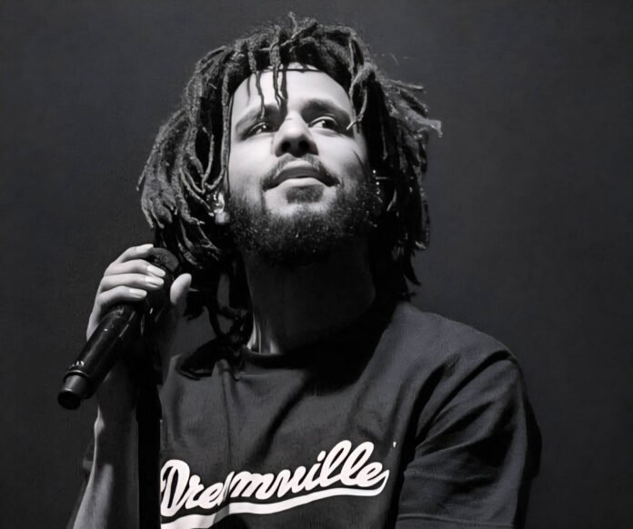 J. Cole performing on stage during a live concert, holding a microphone, wearing a Dreamville t-shirt.