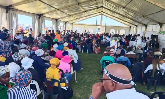 South African social grant beneficiaries gather under a white tent at a SASSA payment point.