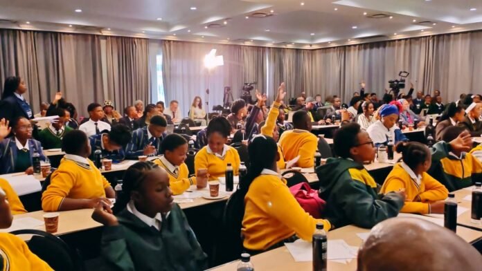 School learners in yellow and green blazers raise their hands at a CDA town hall meeting at the Birchwood Hotel in Boksburg, asking questions about drugs in schools while officials listen.