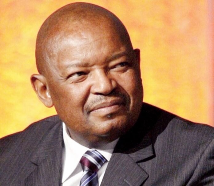 Portrait of Mosiuoa “Terror” Lekota in a dark pinstriped suit and striped tie, shown in a thoughtful close-up with soft lighting and a warm blurred background.