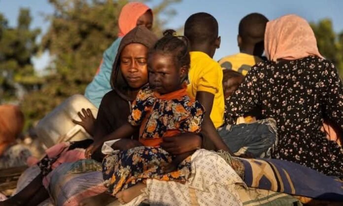 Sudanese children and families displaced by war sit with belongings, reflecting the humanitarian crisis and impact of conflict on civilians.