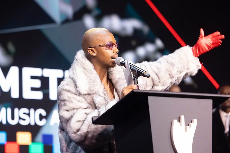 Artist Ciza wearing a large grey fur coat and a red glove while speaking at a podium during the Metro FM Music Awards 2026.