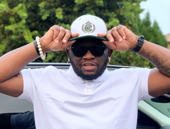 Scooby Nero in white polo and aviator sunglasses adjusting a two-tone cap with green brim and crest.