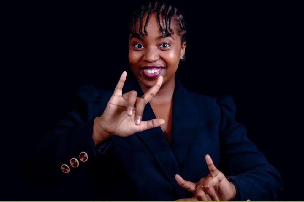 Portrait of Andiswa Precious Gebashe, a South African Sign Language interpreter and Order of Ikhamanga honouree, smiling warmly against a dark background while signing "I love you" with her hands.