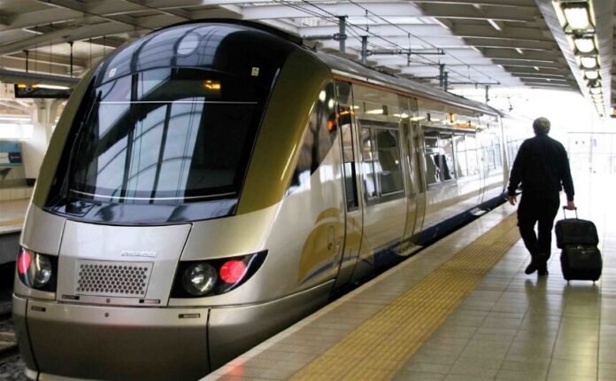 A gold-and-silver Bombardier Gautrain train at a station platform with a commuter walking alongside pulling a black suitcase.