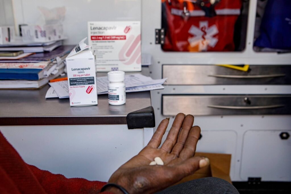 Hand holding Lenacapavir tablets beside HIV prevention medication and injection packaging on a clinic desk