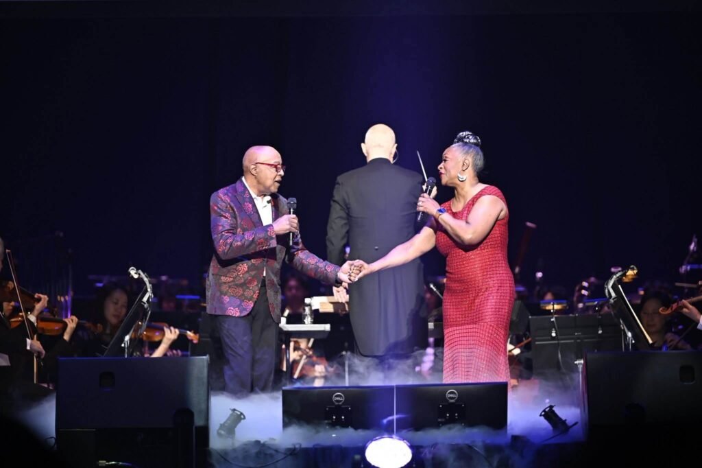 Peabo Bryson and Regina Belle performing a duet on stage with a live orchestra in the background.