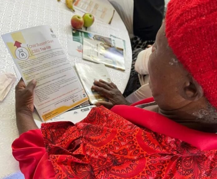 SASSA recipient (mother) holds a Child Support Grant Top-Up flyer at a community table with apples.