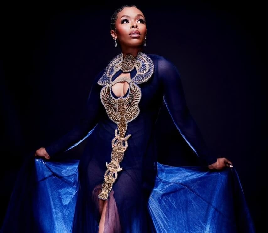 Unathi Nkayi in navy and gold statement collar against dark background, promoting her new single Isikhwele.