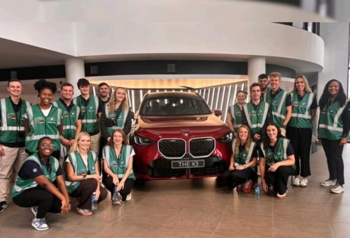 BMW trainees and employees pose in showroom with red BMW X3 during team event at modern dealership.