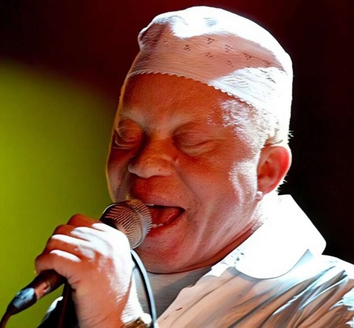 Salif Keita sings into a microphone on stage, eyes closed, under warm stage lighting.