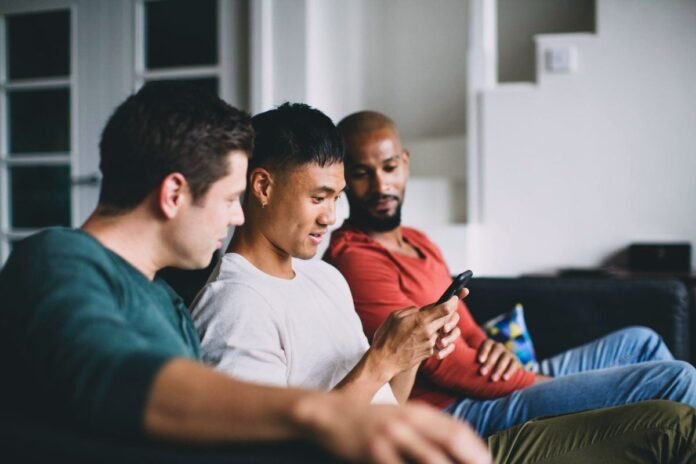 Three men sitting on a couch in a living room and looking at a smartphone together.