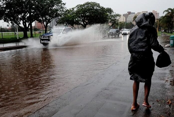 Person in raincoat beside a flooded road as vehicles drive through heavy rain in South Africa.