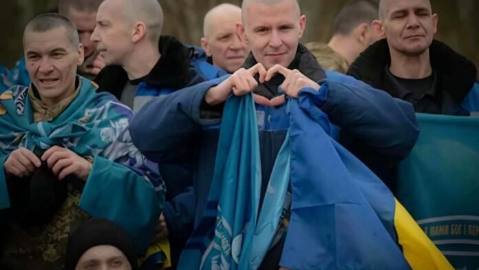 Ukrainian service members return from Russian captivity Released Ukrainian service members sit together, wrapped in Ukrainian flags, as one man forms a heart with his hands.