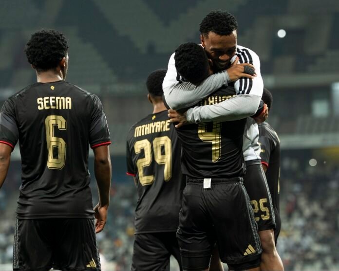 Orlando Pirates players in black kit embrace in celebration on the pitch.
