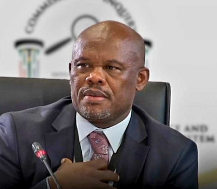 Lt-Gen Shadrack Sibiya in a dark suit seated before a blurred Commission of Inquiry backdrop