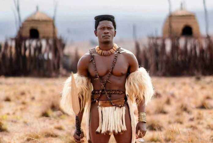 Lemogang Tsipa as King Shaka in Shaka iLembe, holding a spear before thatched huts in a dry grassland