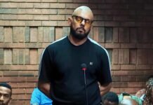 One-way ticket, five bags: Why the State opposes Suliman Carrim’s bail Suliman Carrim stands beside a microphone in court, wearing amber-tinted glasses and a black T-shirt.