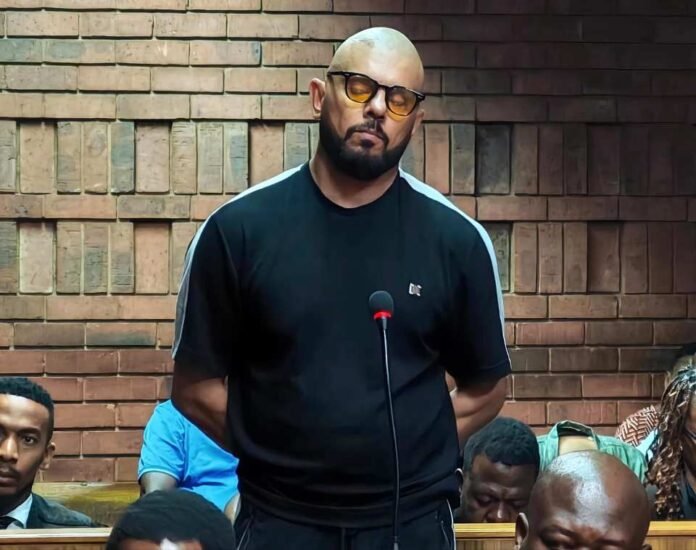 Suliman Carrim stands beside a microphone in court, wearing amber-tinted glasses and a black T-shirt.